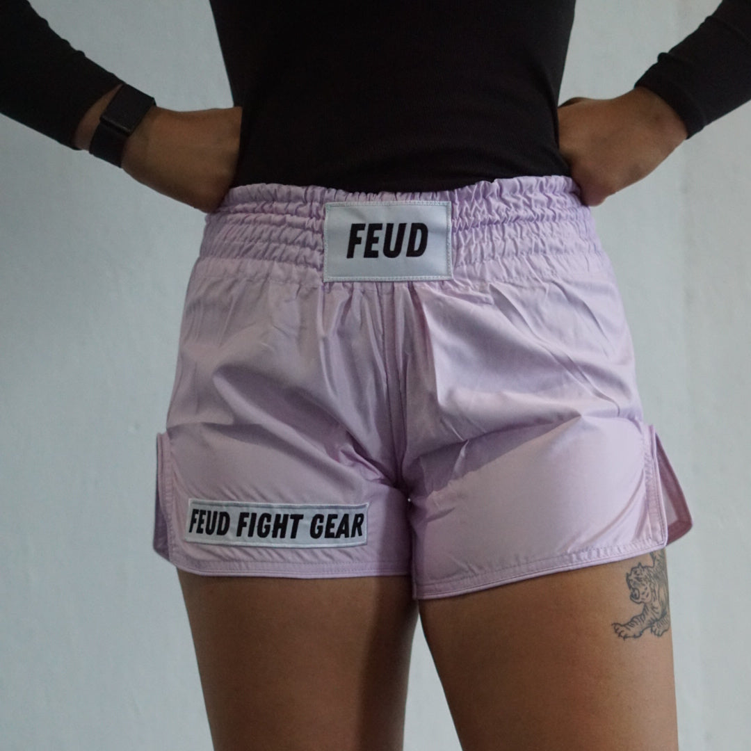 A person wearing lilac Muay Thai shorts with the brand name 'FEUD' on the waistband, standing against a white background.